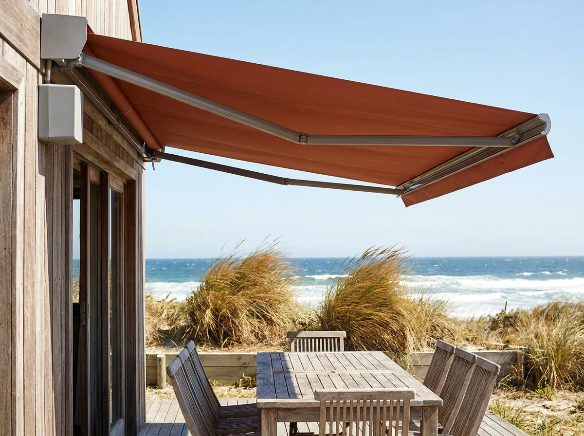 Terracotta folding-arm awning wound out over a Melkbosstrand terrace with a wind sensor mounted beside the cassette, dune grass bending in the background